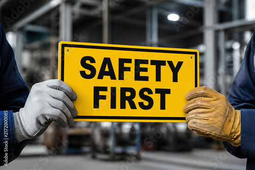 Two people in work attire holding up a bright yellow sign that reads SAFETY FIRST inside an industrial or factory setting.