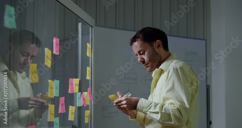 Adult male technology professional writes on sticky notes, compares project ideas and rearranges workflow tasks across glass board during software strategy planning session in modern office.