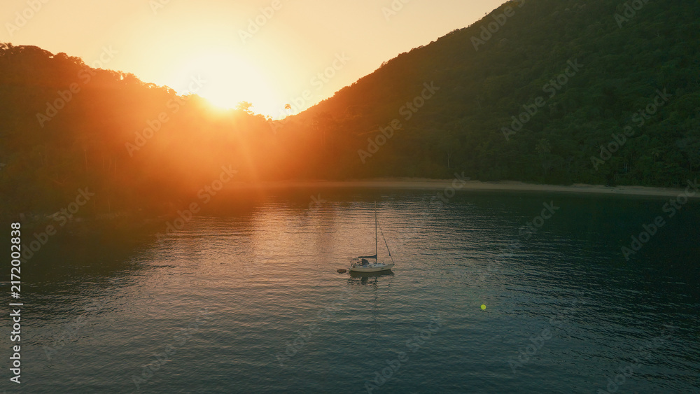 Obraz premium Aerial view of the sailing boat anchored in the calm lagoon with sun rising and shining from the mountains