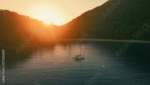 Aerial view of the sailing boat anchored in the calm lagoon with sun rising and shining from the mountains