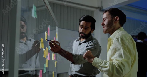 Two adult male technology professionals review sticky notes on glass board, gesture toward project tasks and exchange ideas while discussing software workflow during collaborative strategy session.