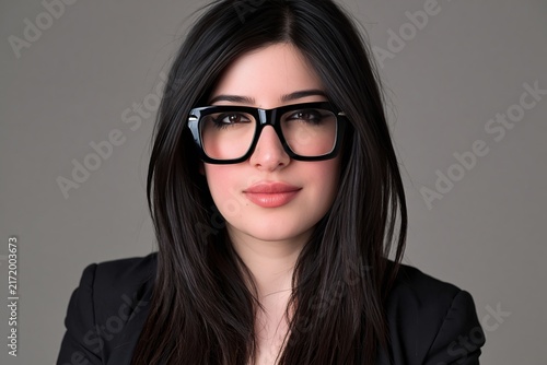Portrait of a confident young woman with dark hair wearing black glasses and a blazer in studio