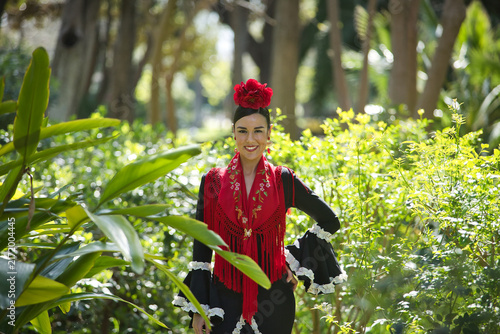 An Andalusian woman wearing a black flamenco dress and a red shawl, with flowers in her hair, posing surrounded by lush greenery that sets off her dress in a park. Concept: dance, art.
