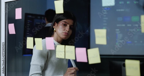 Adult female technology professional writes on sticky notes and places them on glass board, organizing software project ideas and workflow tasks during business strategy planning in modern office.