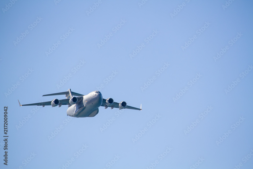 Fototapeta premium C-17 Globemaster III military transport aircraft in flight, viewed from below against a blue sky with a white cloud below the aircraft, showing its four jet engines and distinctive high-wing design.