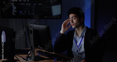 Young Asian man reviews computer monitors while touching temple and face, pausing during demanding software work, supporting workplace stress, system analysis, digital workflow, technology project.