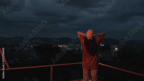 Unrecognizable long-haired man in bucket hat standing on terrace balcony putting hands behind head looking at distant coastal city harbor lights and dark mountains under dramatic night sky rear view