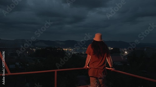 Unrecognizable long-haired person standing on terrace leaning on railing looking at city lights and mountains at twilight back view static shot slow life cozy atmosphere evening lifestyle scenery