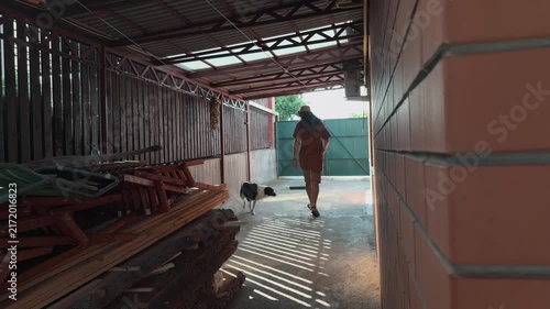 Unrecognizable man with long hair in bucket hat walking away under metal canopy towards green gate in rural backyard with dog and stacked wood planks during sunny summer day authentic rear view