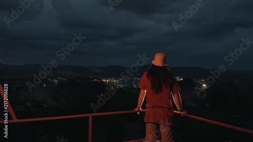 Unrecognizable long-haired person in bucket hat standing on terrace balcony leaning on railing looking at distant coastal city night lights bay port dark mountains under cloudy dusk sky scenery