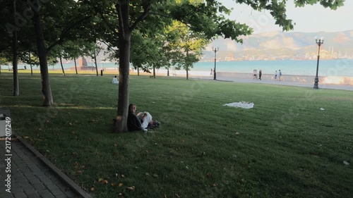 Unrecognizable young woman sitting on green grass under a tree reading a book in coastal city park with pedestrians walking on sea promenade and distant mountains during peaceful summer sunset.