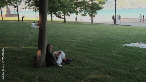Unrecognizable young woman wrapped in blanket sitting under tree on park grass writing in notebook. Creative writer finding inspiration outdoors near sea promenade during golden hour sunset.