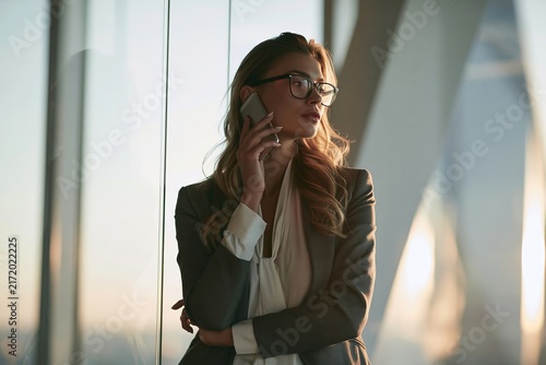Young businesswoman with glasses talking on mobile phone while standing near glass window at sunset