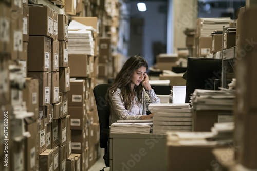 Young woman working at a desk in a cluttered office archive surrounded by many cardboard boxes