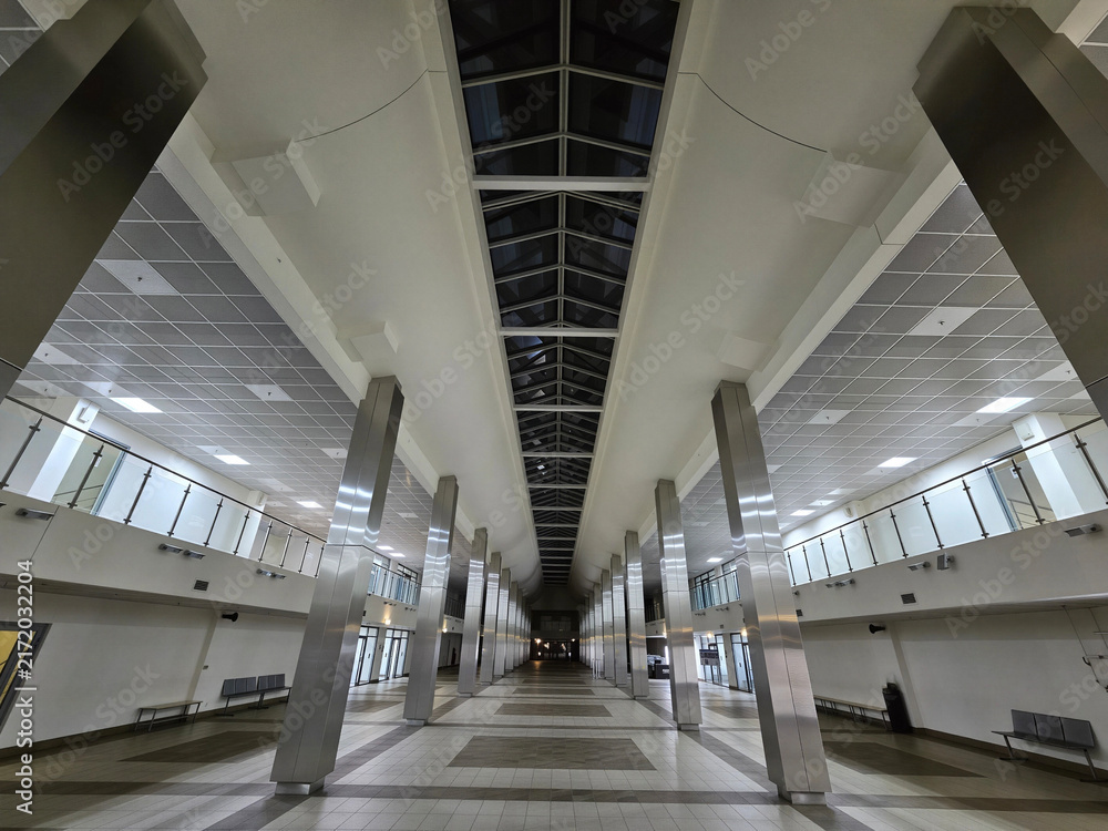 Fototapeta premium Empty station hall with tall columns and skylights. Ceiling panels and tiled floors reflect overhead lighting. Passenger benches line both sides of the corridor