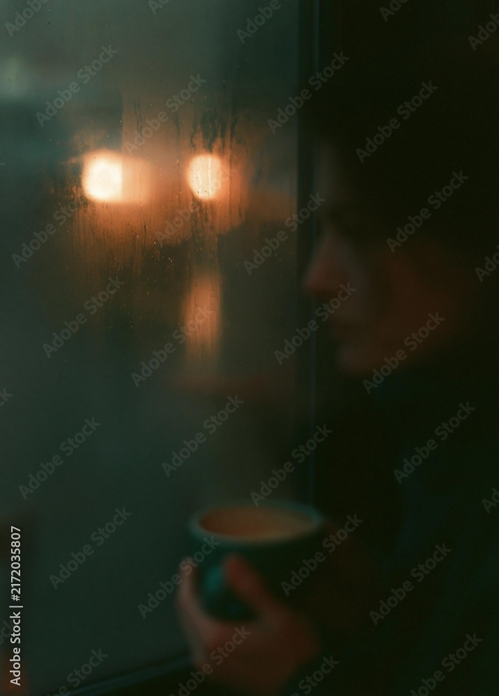 Obraz premium Close-up of a person's face, holding a cup of coffee in their hand. the person is standing in front of a window, and the background is blurred, but it appears to be a cityscape at night.