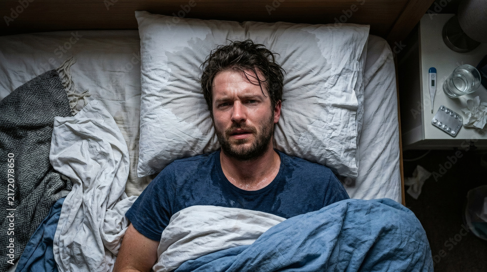 Fototapeta premium Sick Man Lying in Bed With Fever and Sweaty Shirt Surrounded by Tissues and Medicine