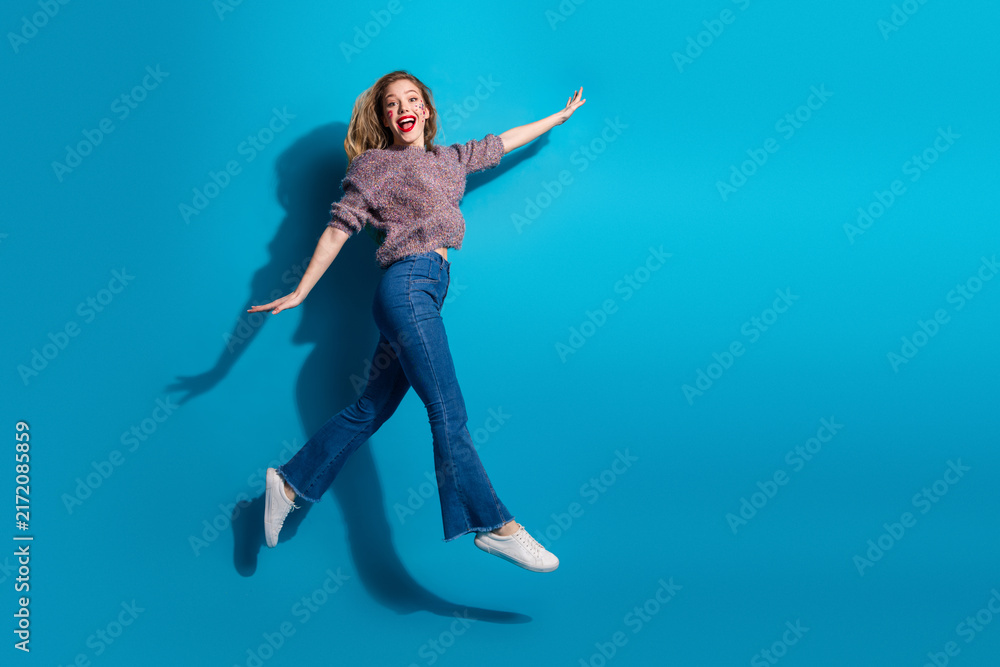 Fototapeta premium Young woman jumping in a blue studio background in casual sweater and jeans showcasing happiness fashion and lifestyle vibe