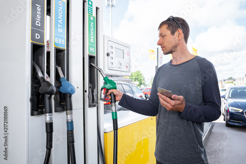 Konstfotografi A man holds a green fuel nozzle and a smartphone while refueling his vehicle at a gas station with diesel, ethanol, and gasoline pumps visible outdoors