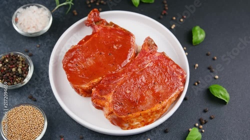 Two marinated pork chops on a white plate with spices and herbs on dark background