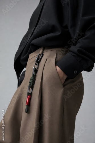 Close-up of urban streetwear outfit featuring olive-green pleated trousers with black jacket, secured with green paracord carabiner, minimalist urban fashion styling, moody lighting
