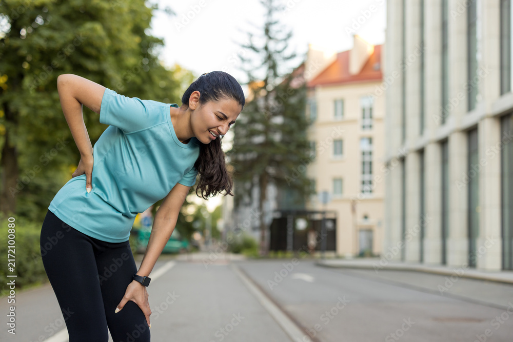 Naklejka premium Woman suffering from acute lower back pain and knee discomfort while exercising on a city street, indicating a sports injury or physical strain during her outdoor fitness routine