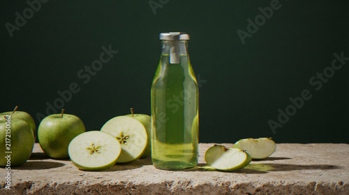 Fotografie A bottle of fresh green apple juice standing on a rustic stone surface surrounded by whole and sliced green apples against a dark background