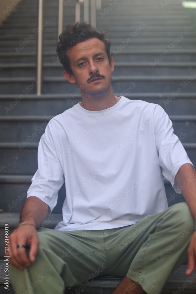 Fototapeta premium Blank white oversized t-shirt mockup on a young male model sitting on city stairs, urban street style apparel template