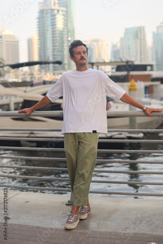 Blank white oversized t-shirt mockup on a young male model, modern city marina background with yachts and skyscrapers