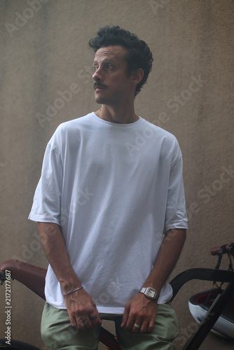 Blank white oversized t-shirt mockup on a young male model sitting on an electric bicycle, moody urban aesthetic