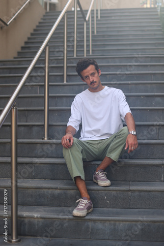 Blank white oversized t-shirt mockup on a young male model sitting on city stairs, urban street style apparel template