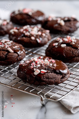 Warm, freshly baked chocolate cookies sit cooling on a wire rack, adorned with crushed peppermint candy. Their glossy chocolate glaze sparkles under soft light, inviting a taste of joy