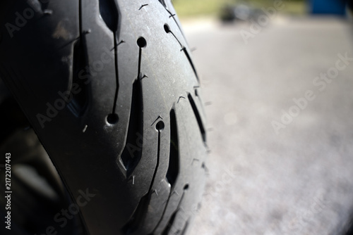 Close-up of a motorcycle tire with deep treads. Details highlight texture and wear on the rubber surface. Tire features multiple circular punctures and sharp grooves