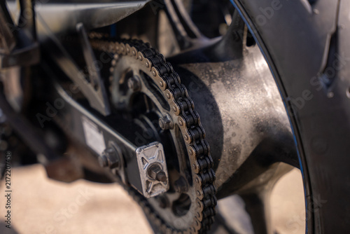 Motorcycle chain links grip metallic sprocket tightly. Rust and grime coat exposed gear assembly. Shallow depth of field highlights mechanical detail. Industrial grit suggests rugged