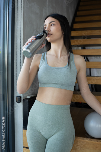 Young woman in athletic wear drinks water from a reusable bottle after workout