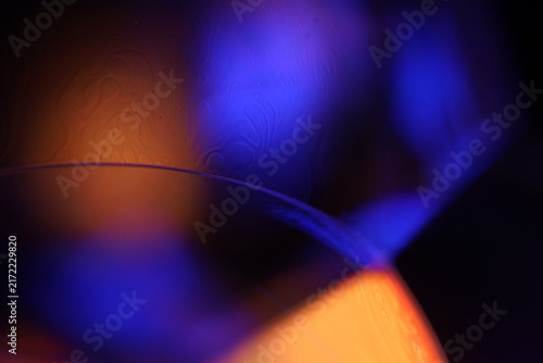 Thin fluid edge arc with vibrant blue and warm amber bokeh backlighting. A razor-thin specular arc traces a liquid membrane edge in low-key macro perspective.