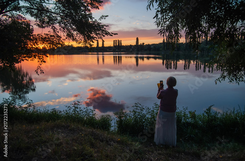 Woman photographing a tranquil lake at sunset, with colorful reflections, silhouetted trees, and a peaceful summer landscape.