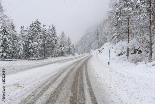 Snow covered road through a forested landscape in the European Alps during a heavy snowfall in winter. Driver point of view.