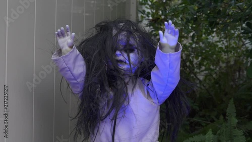 Spooky doll prop with a pale face, messy black hair, and raised hands in a yard. Halloween outdoor decoration in a garden with green plants creating a horror mood.