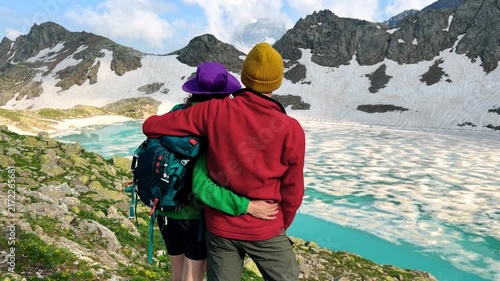 Couple Embracing While Hiking Above a Turquoise Alpine Lake Surrounded by Snowy Mountains