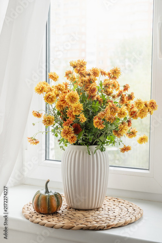 Orange chrysanthemum bouquet in white vase with small pumpkin on bright windowsill, seasonal autumn home decor with natural daylight. Vertical format. Cozy home interior