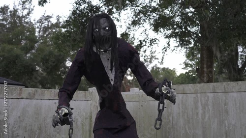 Tall spooky ghoul with glowing eyes holds chains by a weathered white fence. Halloween yard decoration in a suburban outdoor lawn creating a scary horror scene.