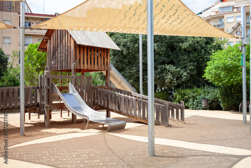 Wooden playground structure with metal slide under large shade sails in a public urban park. Outdoor recreational area for children with safe play equipment, natural materials and sunny day atmosphere