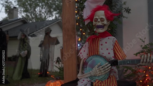 Close-up of a skeleton clown with glowing red eyes playing a banjo at dusk. Orange lights, a pumpkin, and witches in the background create a spooky Halloween yard decoration.