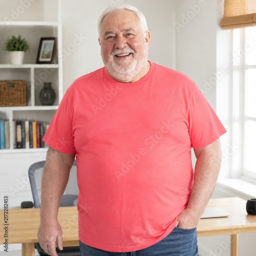 Cheerful older man in a coral shirt smiling at the camera in a bright, cozy home office setting.