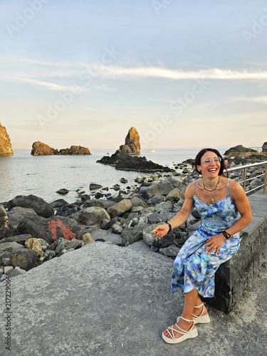 Smiling brunette woman wearing blue sundress and glasses rests on promenade wall overlooking the Cyclopean Isles with iconic volcanic sea stacks on a summer evening in Aci Trezza, Sicily, Italy.