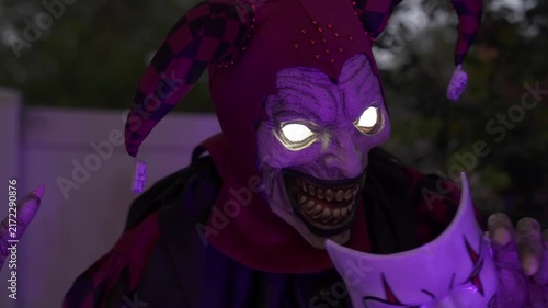 Close-up of a scary Halloween jester clown with glowing eyes at night. The figure holds a white mask, showing a creepy evil smile as an outdoor yard decoration.