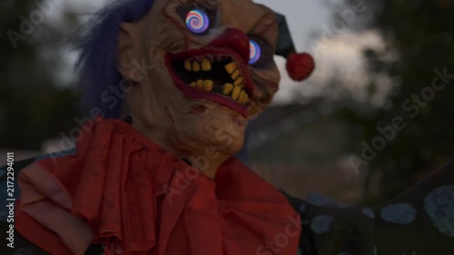 Close-up of a scary Halloween clown with spiral glowing eyes, green face, rotten yellow teeth, blue hair, and a red nose at night. Outdoor yard decoration with an orange collar creates a spooky horror