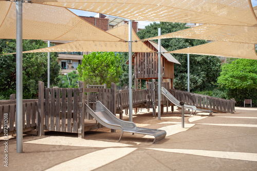 Wooden playground structure with metal slide under large shade sails in a public urban park in Barcelona, Spain. Outdoor recreational area for children with safe play equipment, natural materials 