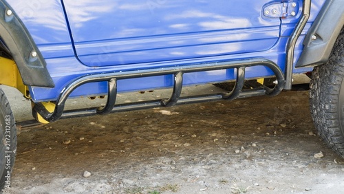 Close up view of black rock slider bar under blue car door panel with visible yellow leaf spring suspension.